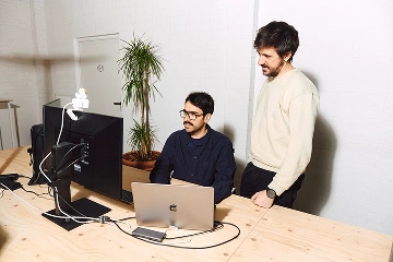 Bernat i Andreu col·laborant en un escriptori amb monitors i un portàtil